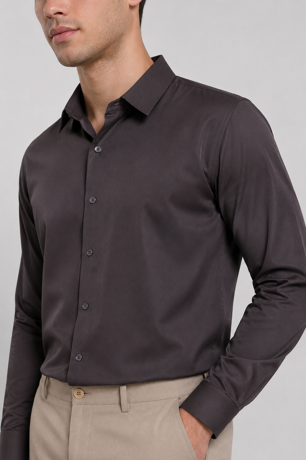 A man wearing a dark charcoal gray long-sleeve button-down shirt and beige trousers, shown from the chin down against a solid light gray background.