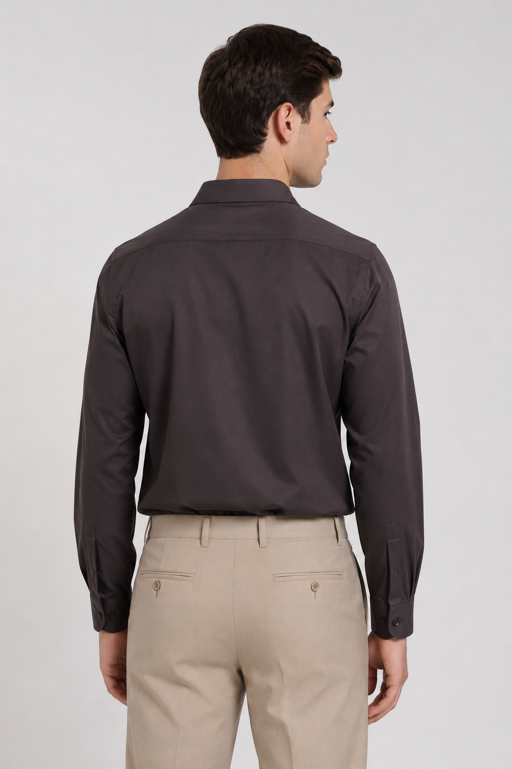 A man shown from the back wearing a dark charcoal gray long-sleeve button-down shirt tucked into light beige trousers, against a light gray studio background.